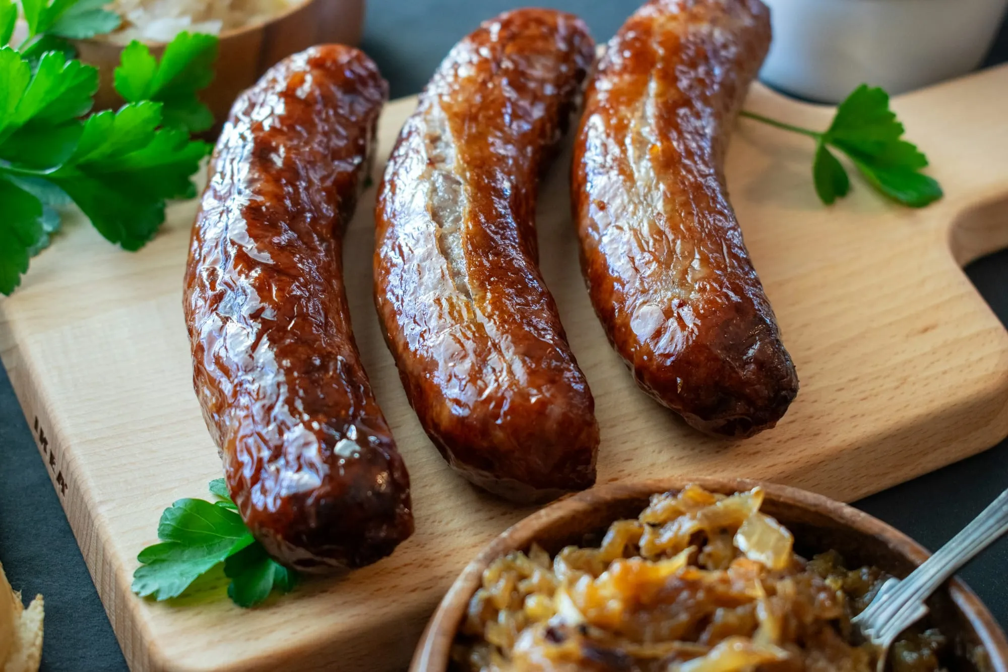 Three browned bratwursts on a wooden board with a bowl of caramelized onions (stock photo)