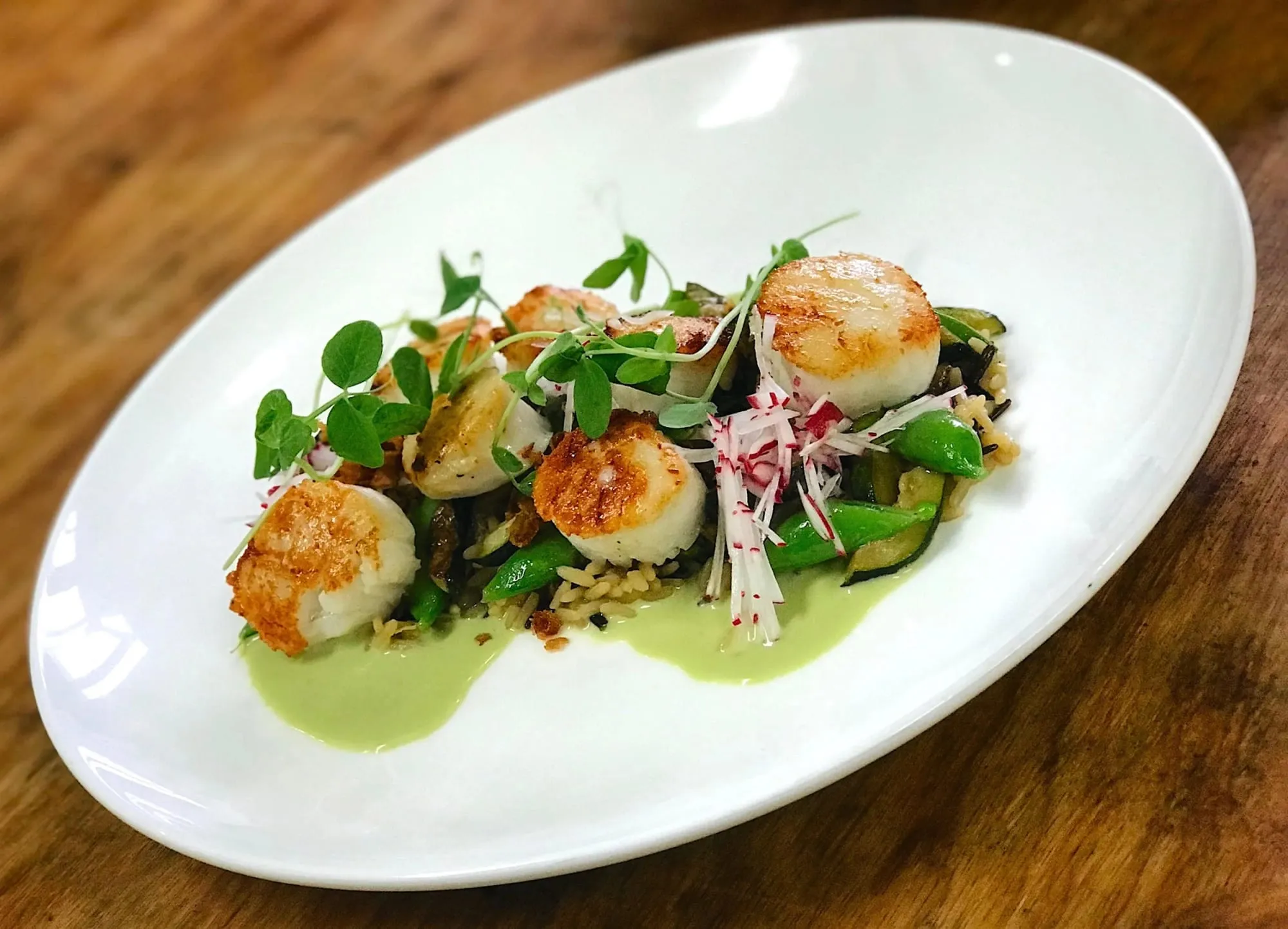 Seared scallops over green vegetables and a pale green sauce in a white oval dish