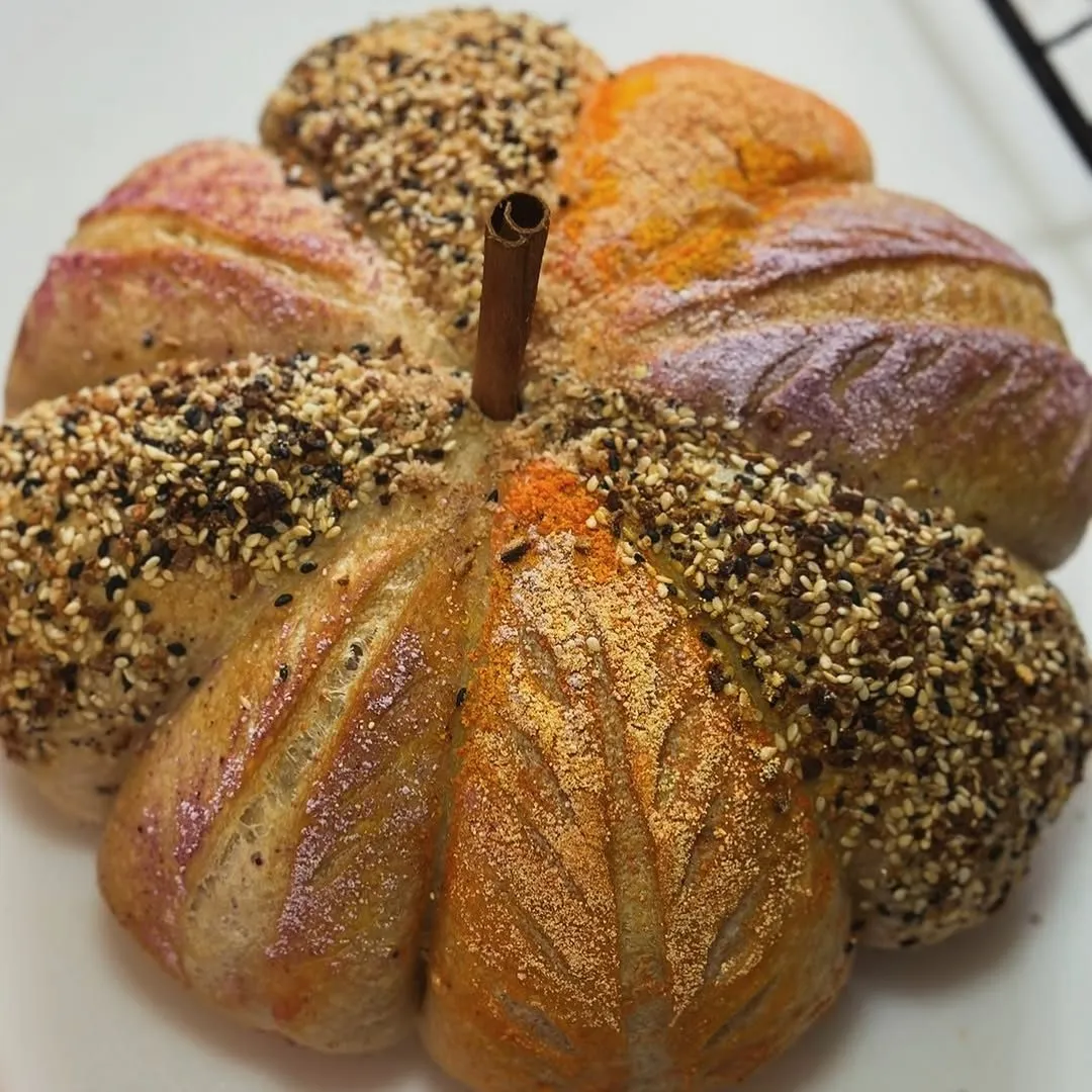 A pumpkin-shaped sourdough loaf with a cinnamon stick stem, its sections topped with sesame seeds and dusted with spices