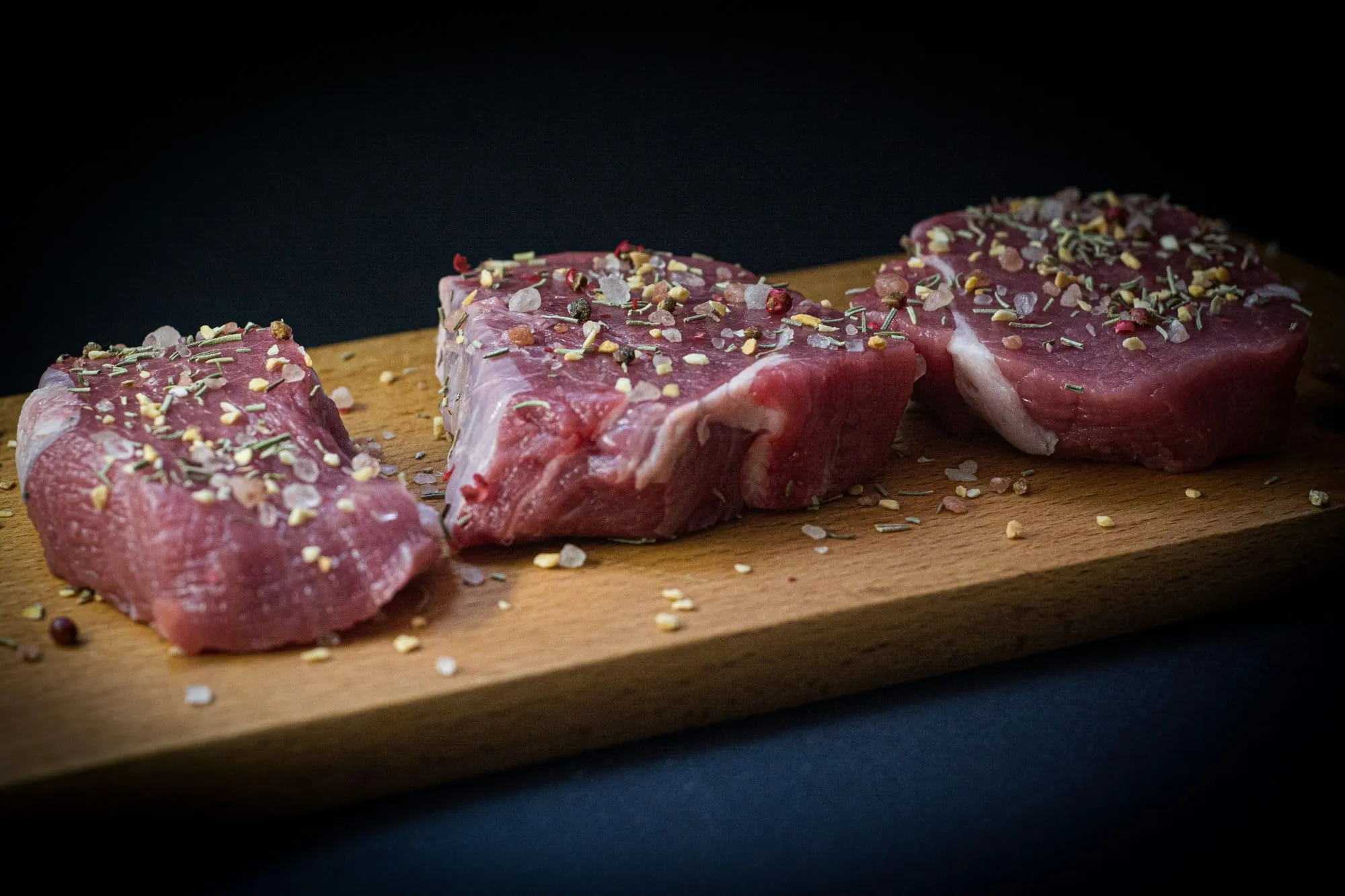 Thick raw beef steaks seasoned with herbs on a wooden board against a dark background (stock photo)