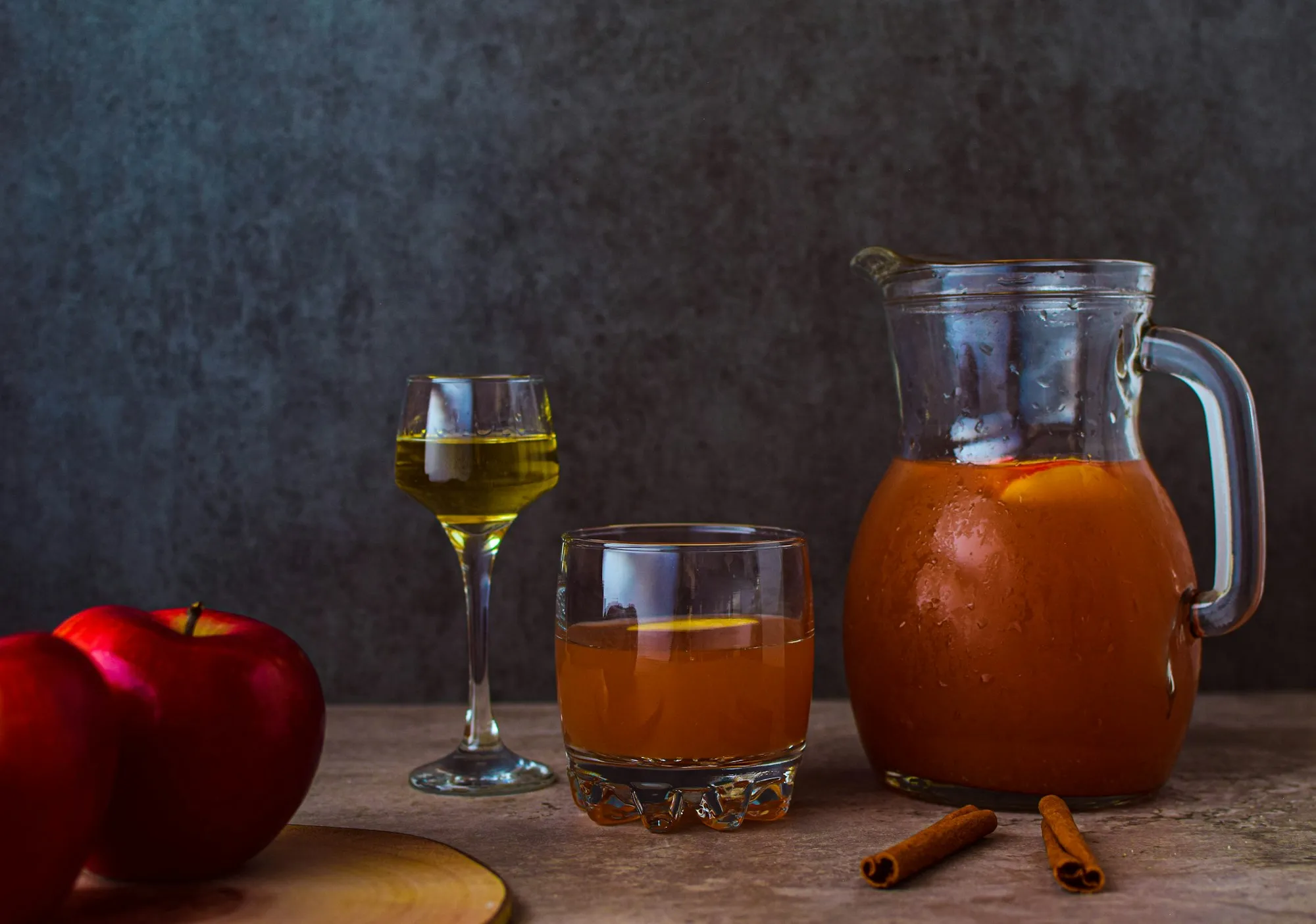 A glass pitcher and glasses of amber apple cider beside red apples and cinnamon sticks (stock photo)