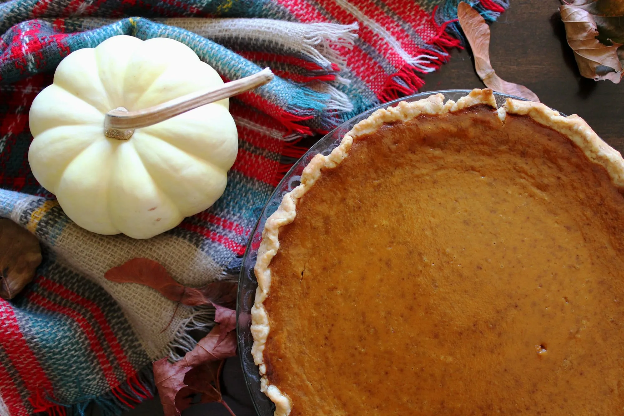 A whole pumpkin pie with a fluted crust beside a small white pumpkin on a plaid blanket (stock photo)