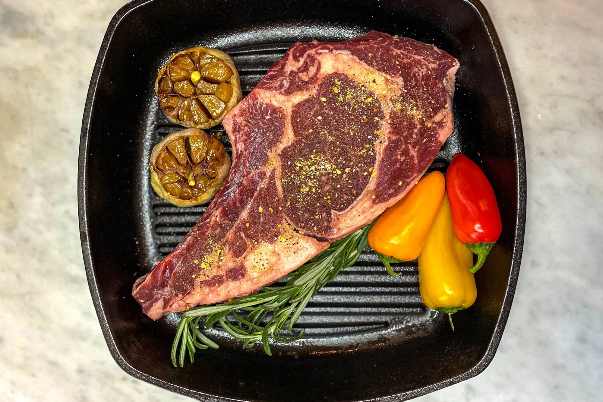 A seasoned bone-in ribeye in a cast iron grill pan with roasted garlic, rosemary and sweet peppers