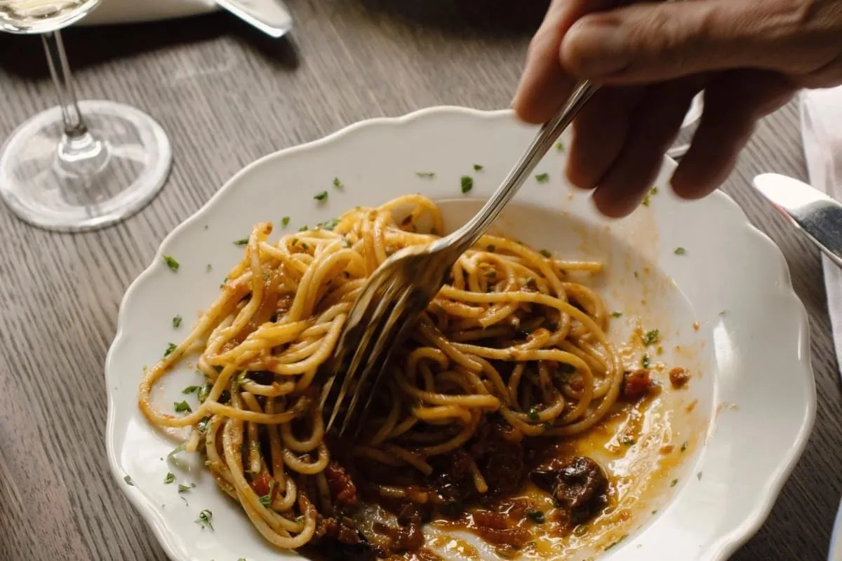 A fork twirling spaghetti in a scalloped bowl on a wooden table beside wine glasses