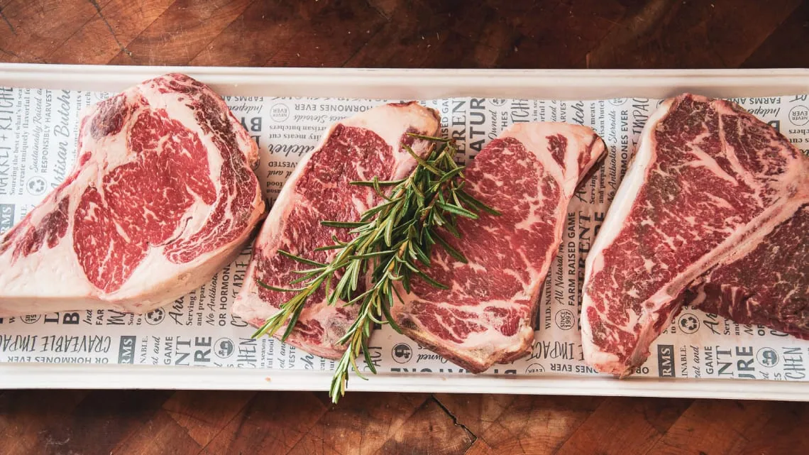Four raw marbled bone-in steaks with a sprig of rosemary on a butcher-paper-lined tray