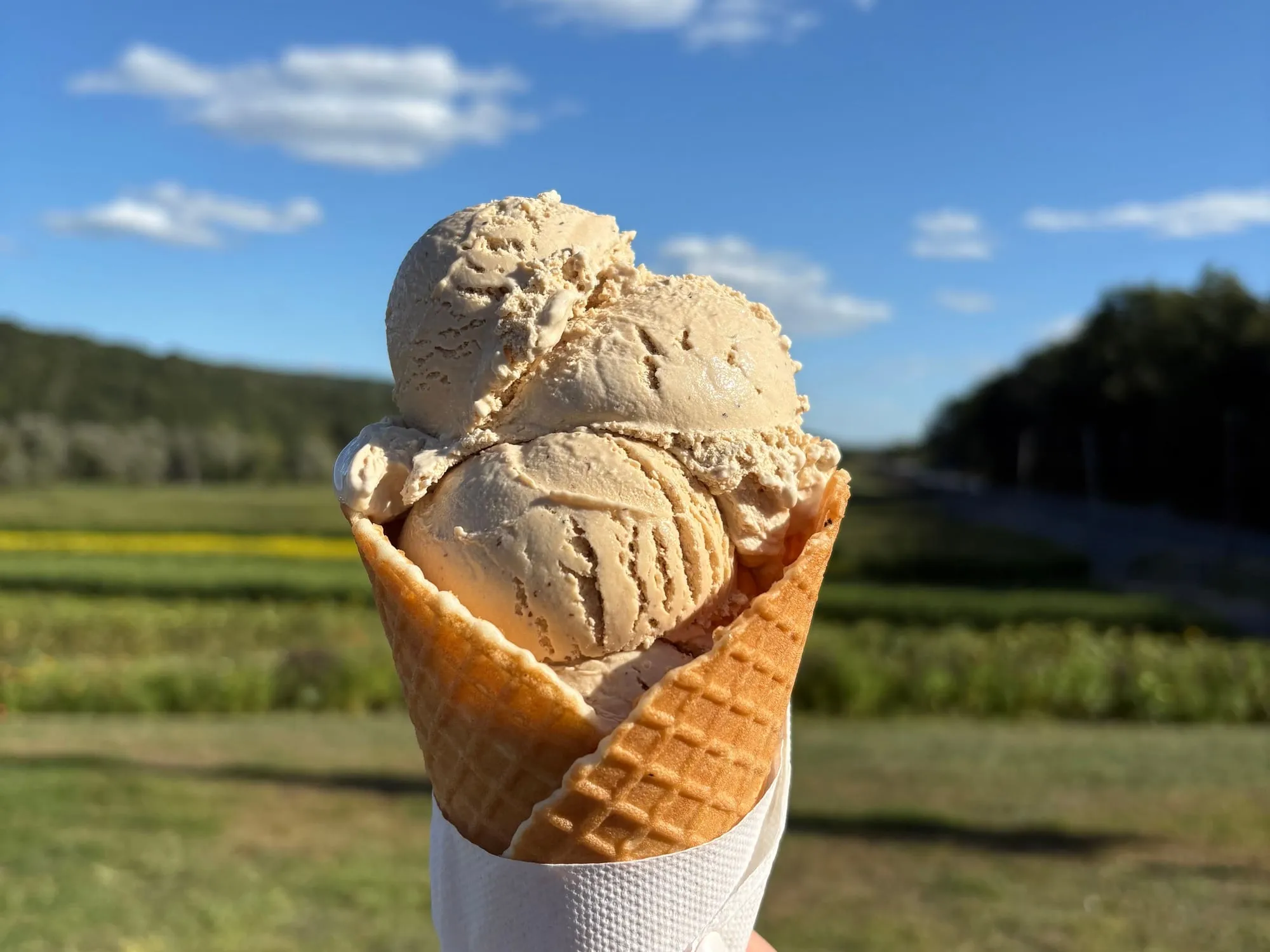 A triple scoop of homemade coffee-colored ice cream in a waffle cone against the farm fields