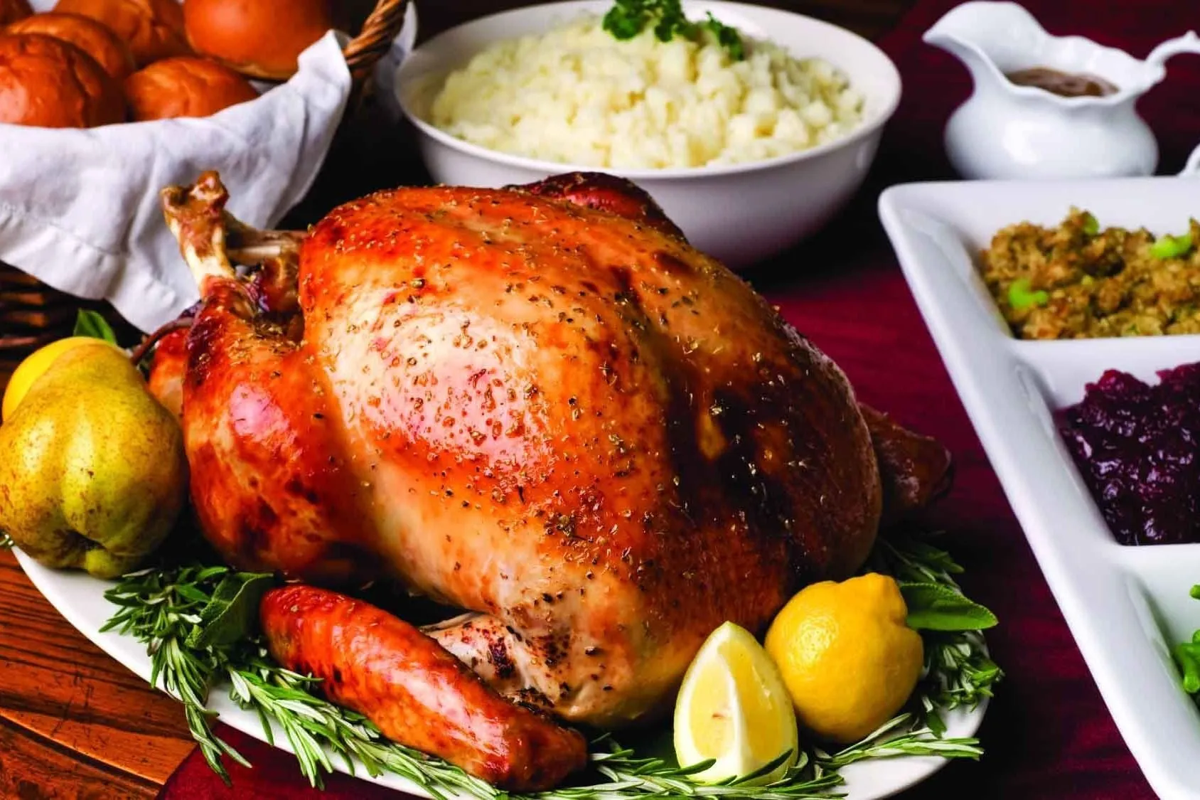 A roast turkey on a platter with rosemary, lemons and pears, beside mashed potatoes, stuffing and cranberry sauce