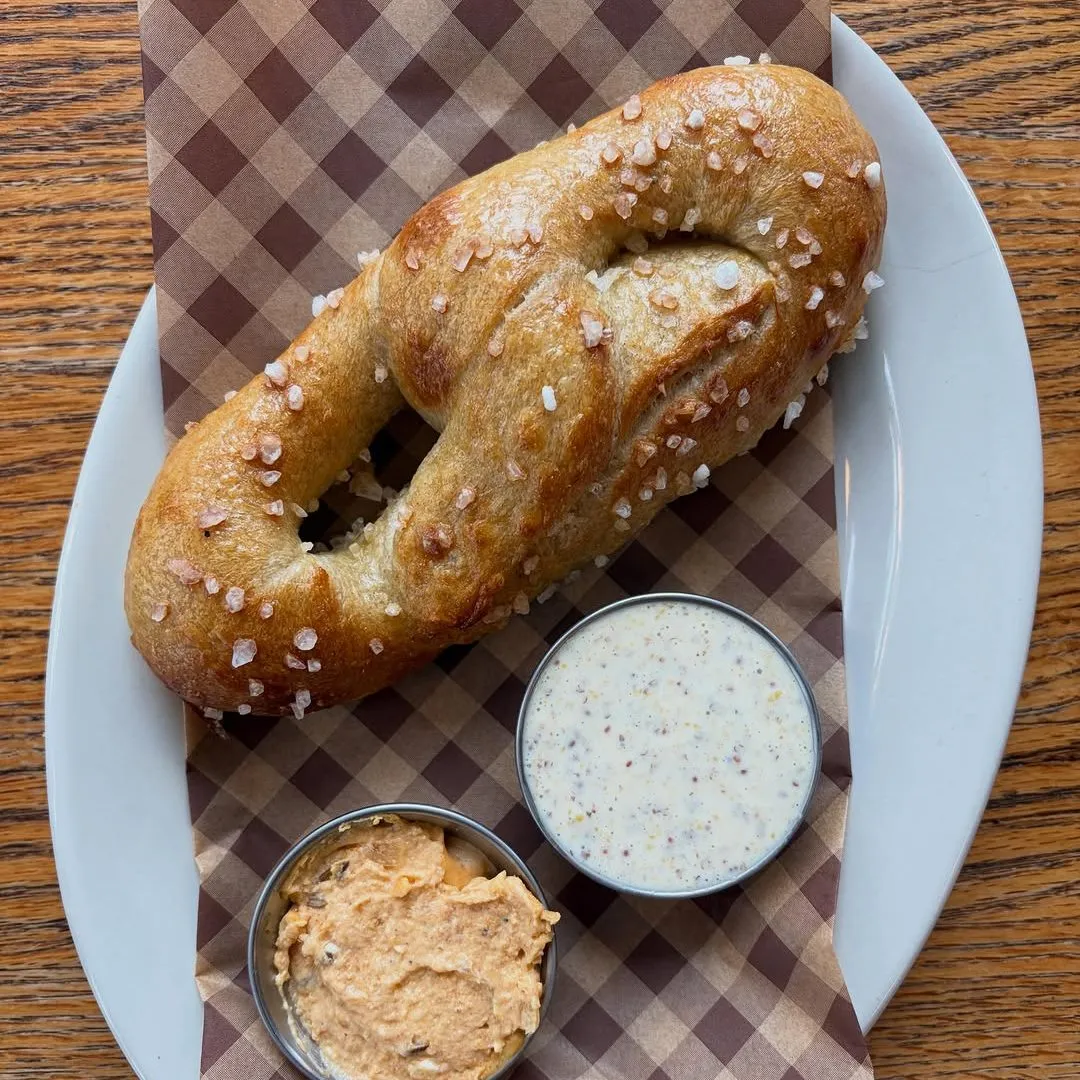 A salted sourdough pretzel on checked paper with two small cups of dip