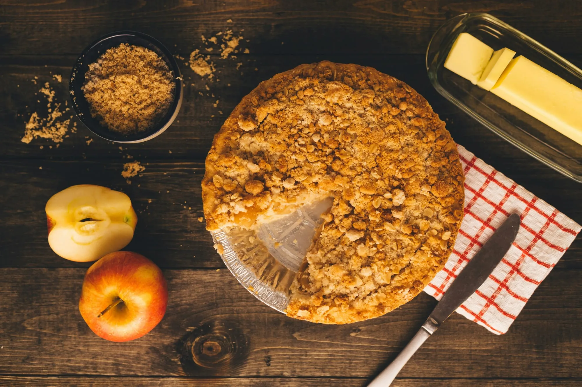 A Dutch apple crumb pie with a slice cut, beside fresh apples and butter (stock photo)