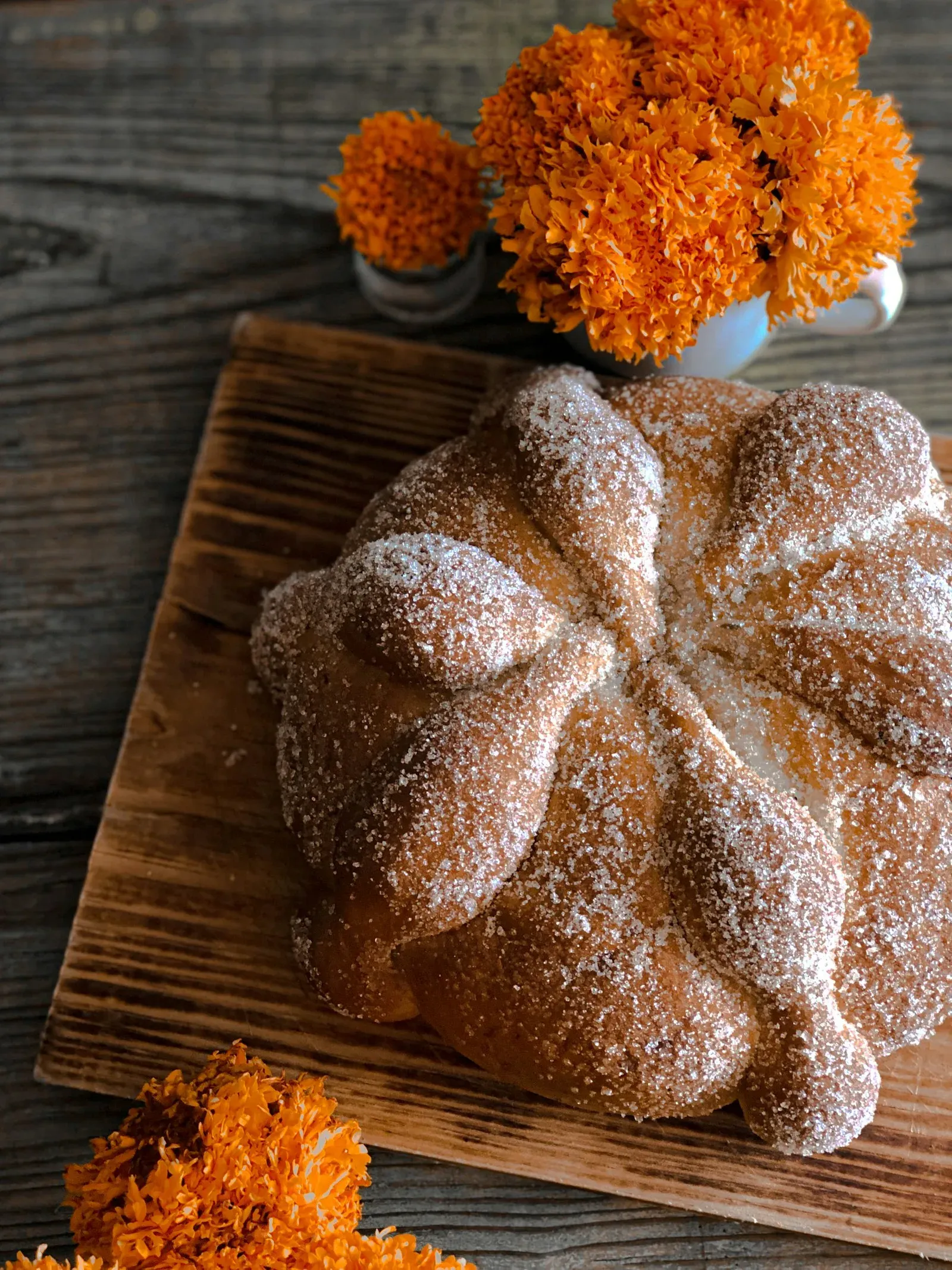 A sugar-crusted pan de muerto on a wooden board with orange marigolds