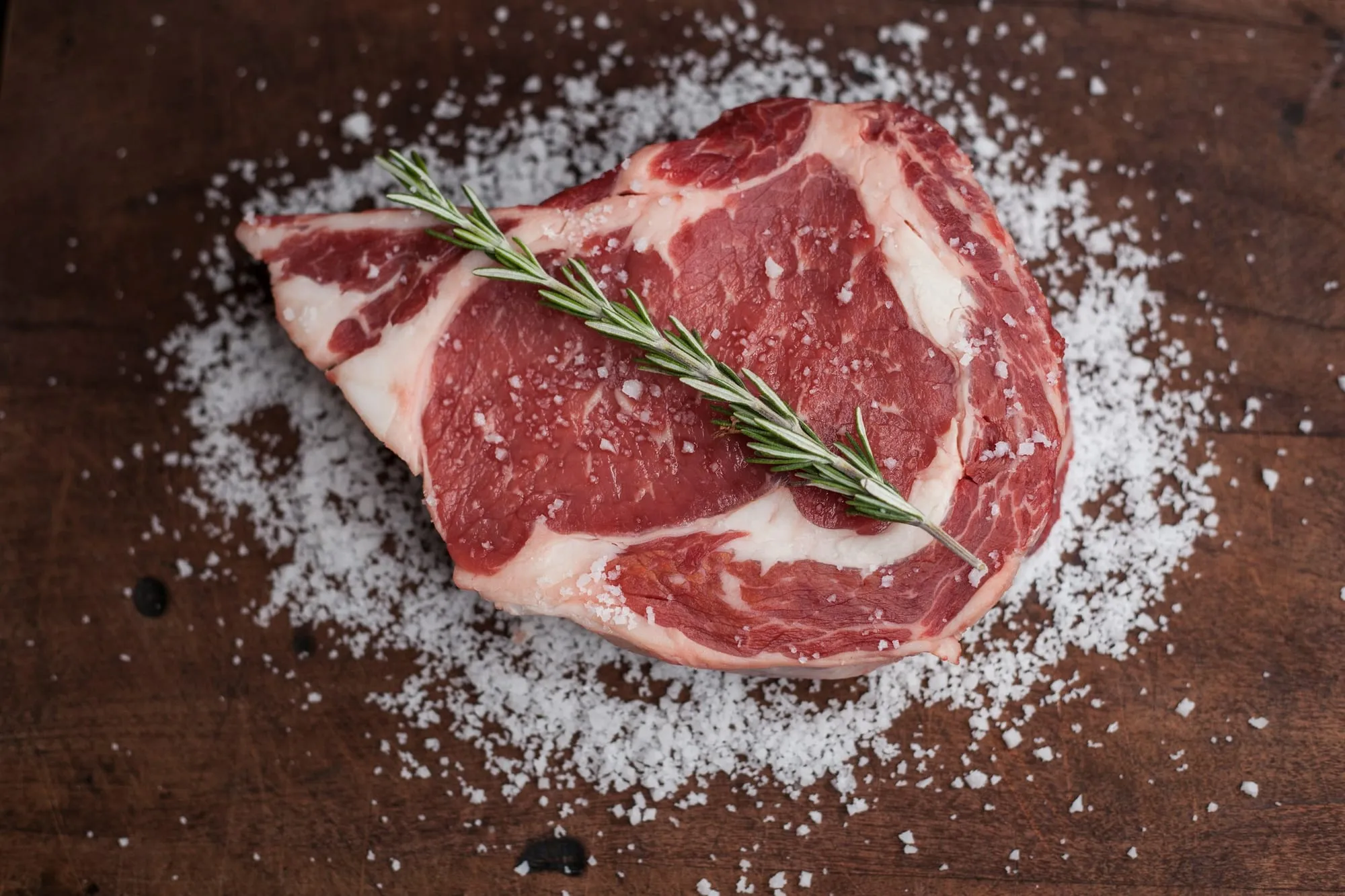 A raw bone-in ribeye steak with rosemary on coarse salt (stock photo)