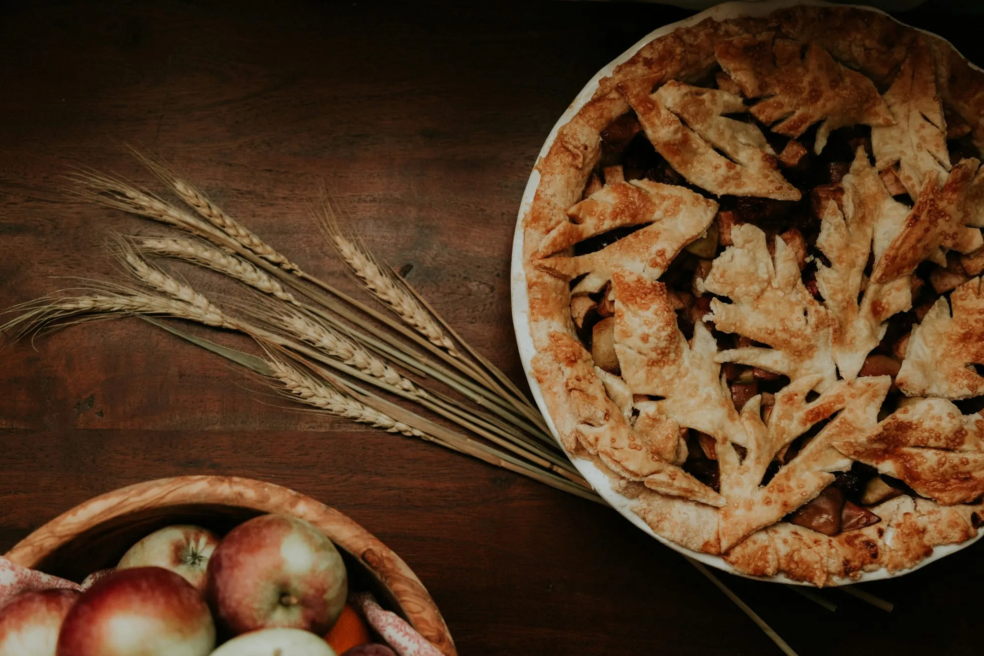 An apple pie with a leaf-cut crust beside a bowl of apples and wheat stalks (stock photo)