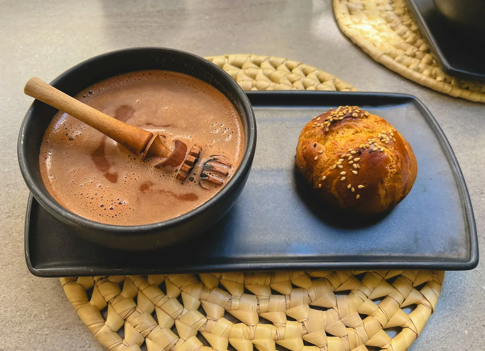 A sesame pan de muerto beside a bowl of hot chocolate with a wooden molinillo