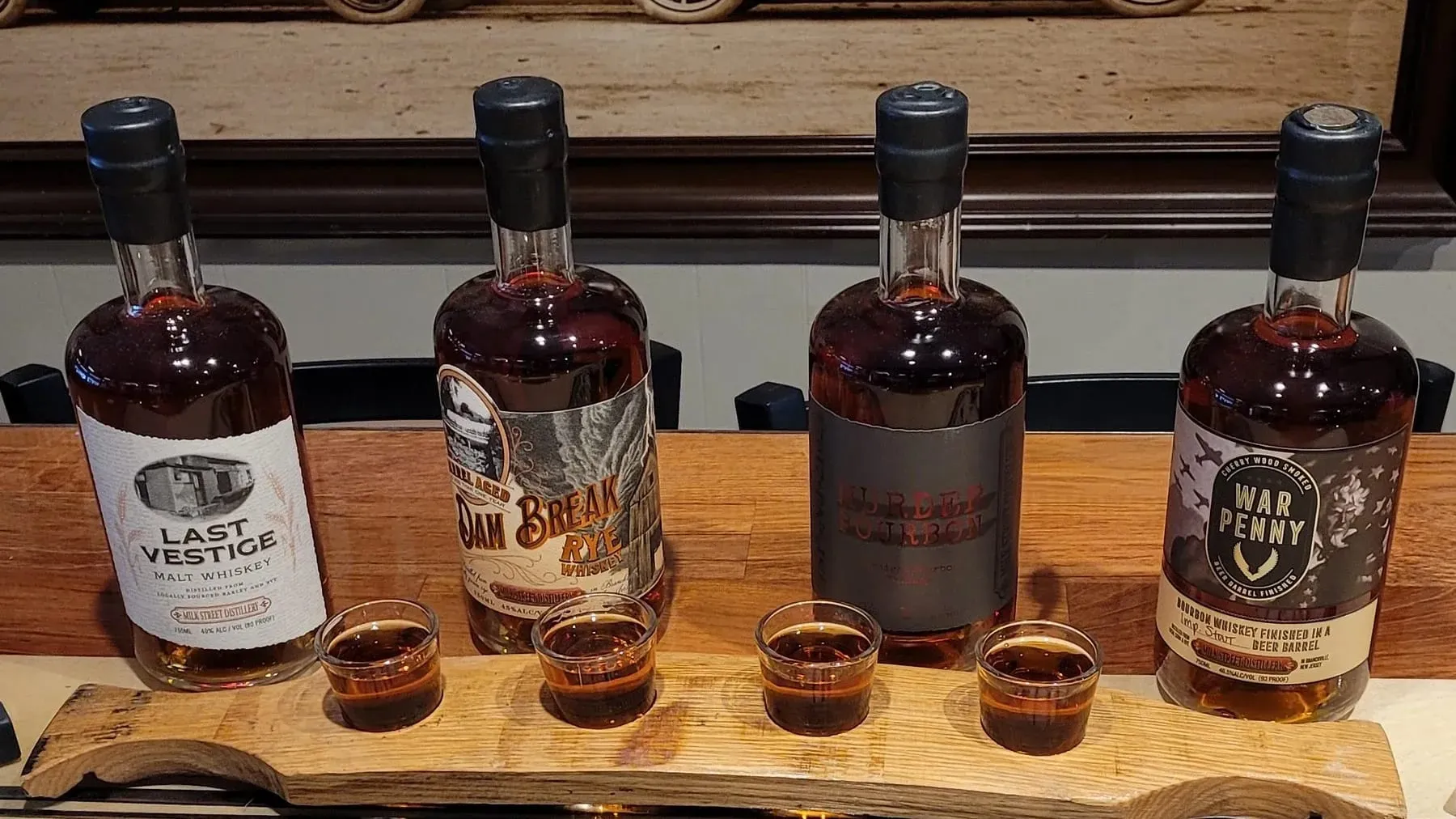 A whiskey tasting flight on a wooden paddle in front of four Milk Street Distillery bottles