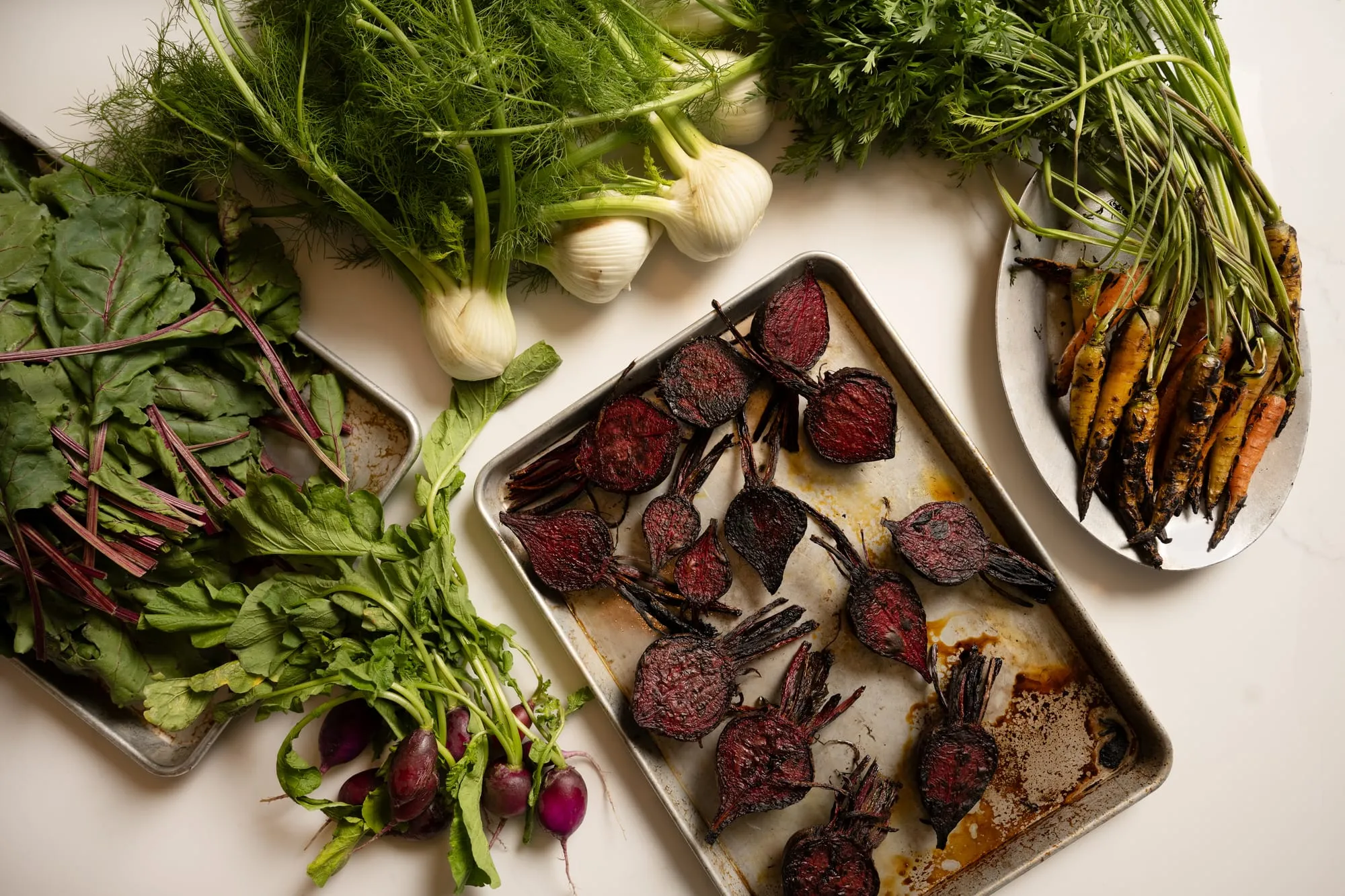 Roasted beets on a sheet pan beside bunches of fennel, beet greens, radishes and charred carrots from the farm