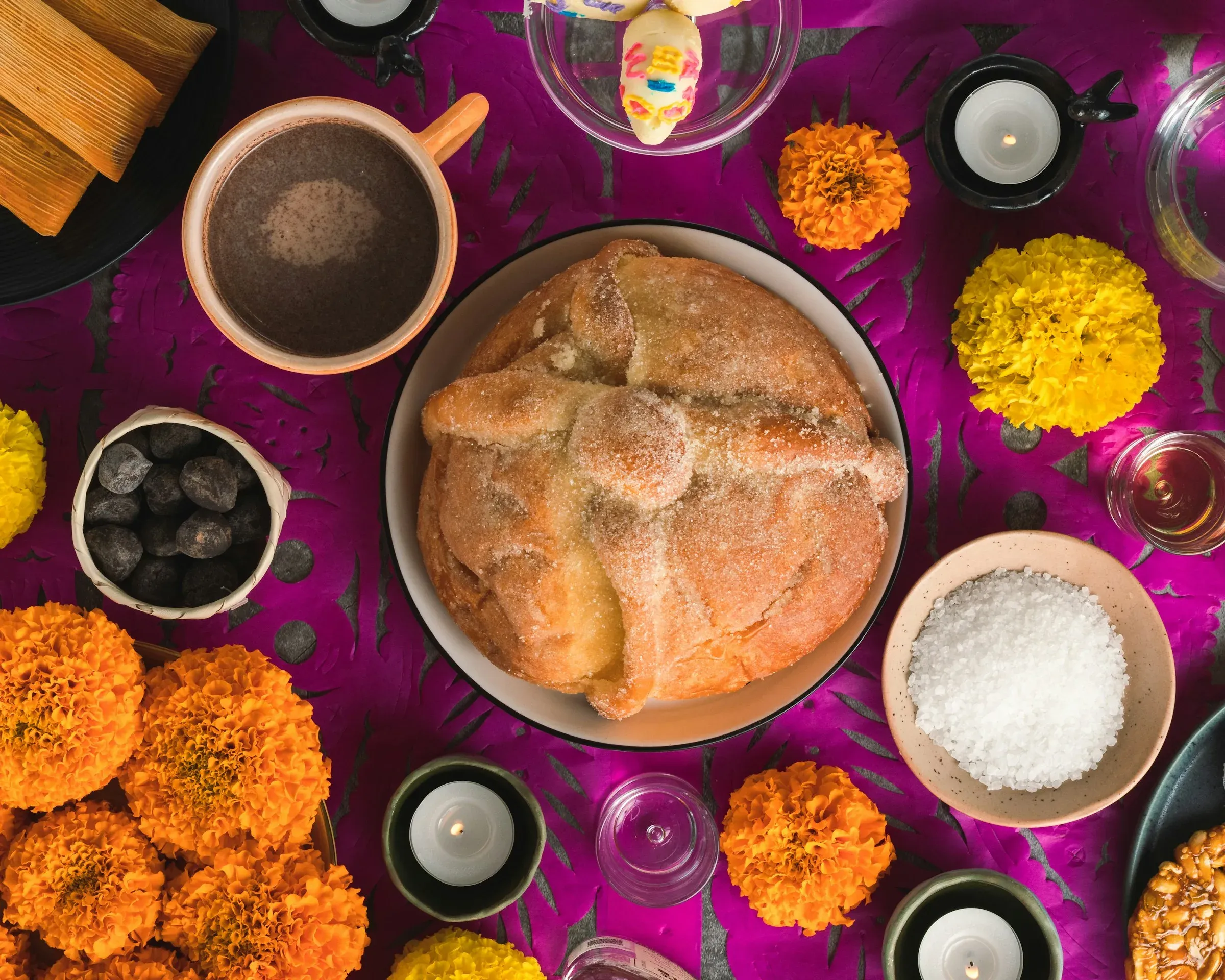 A round pan de muerto on an ofrenda table with marigolds, candles, hot chocolate and tamales