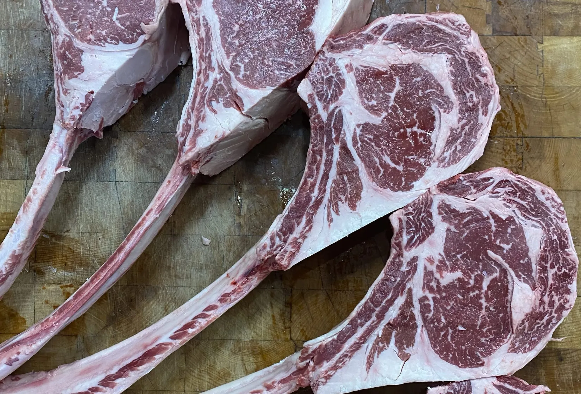 Four raw tomahawk ribeye steaks with long frenched bones on a butcher block