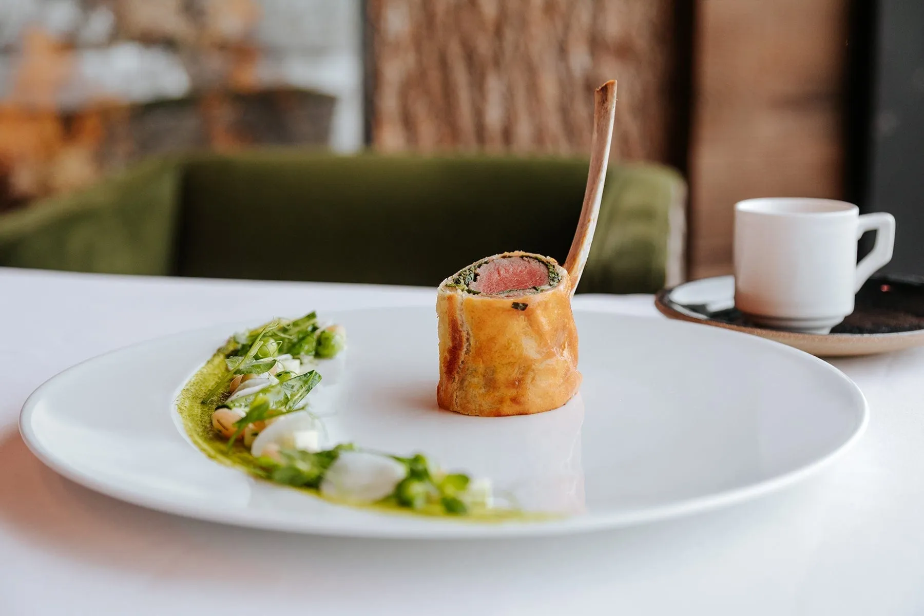 A slice of meat wellington with a bone handle on a white plate with green sauce and spring vegetables