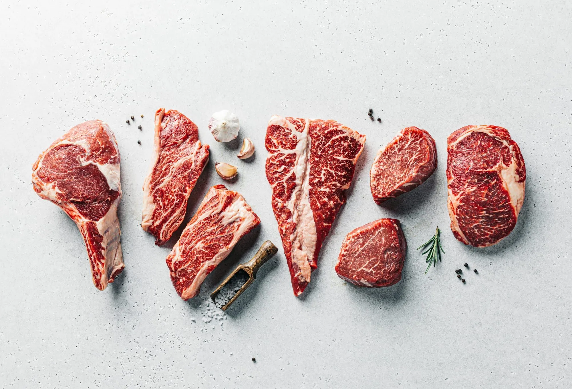 Marbled raw beef steaks and chops laid out on pale stone with garlic, peppercorns and rosemary (stock photo)