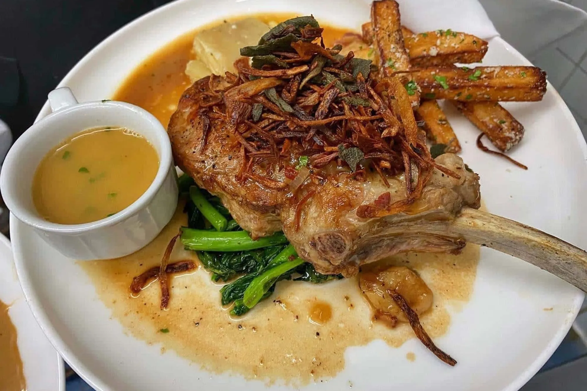 A roasted bone-in pork chop with crispy shallots, carrots and broccoli rabe beside a cup of sauce