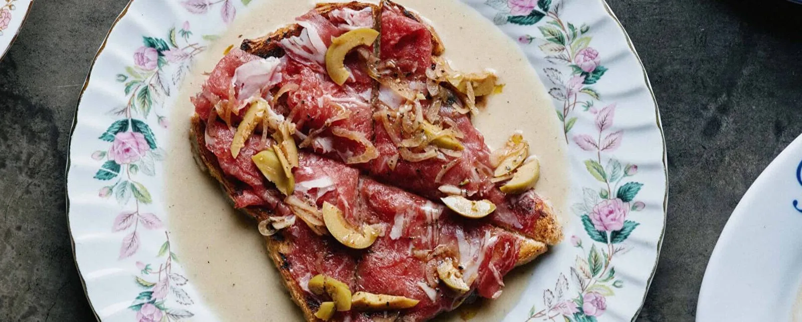 Beef carpaccio with olives and caramelized onion on grilled toast, on a floral china plate