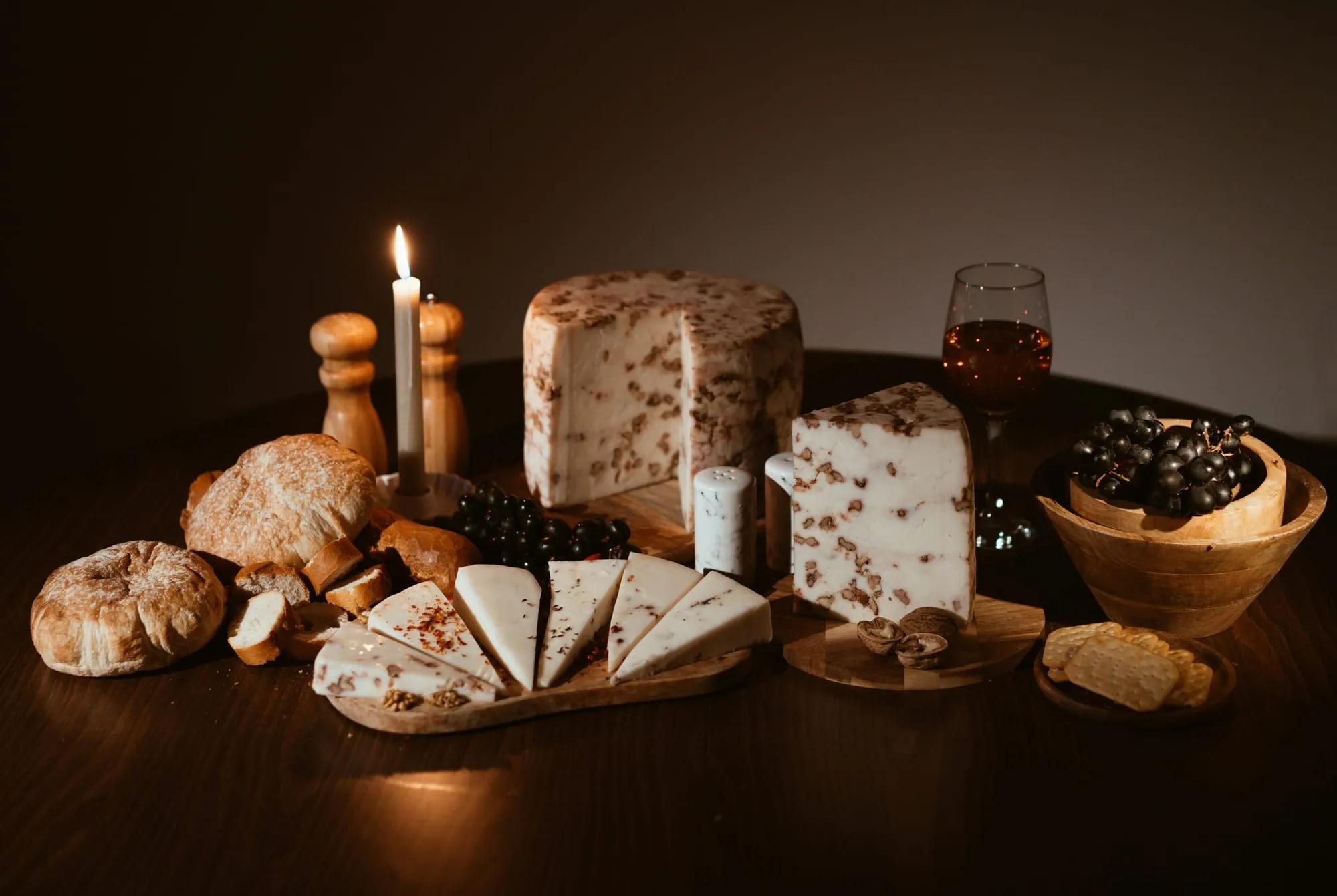 Wheels and wedges of farmstead cheese with bread and candlelight on a wooden table (stock photo)