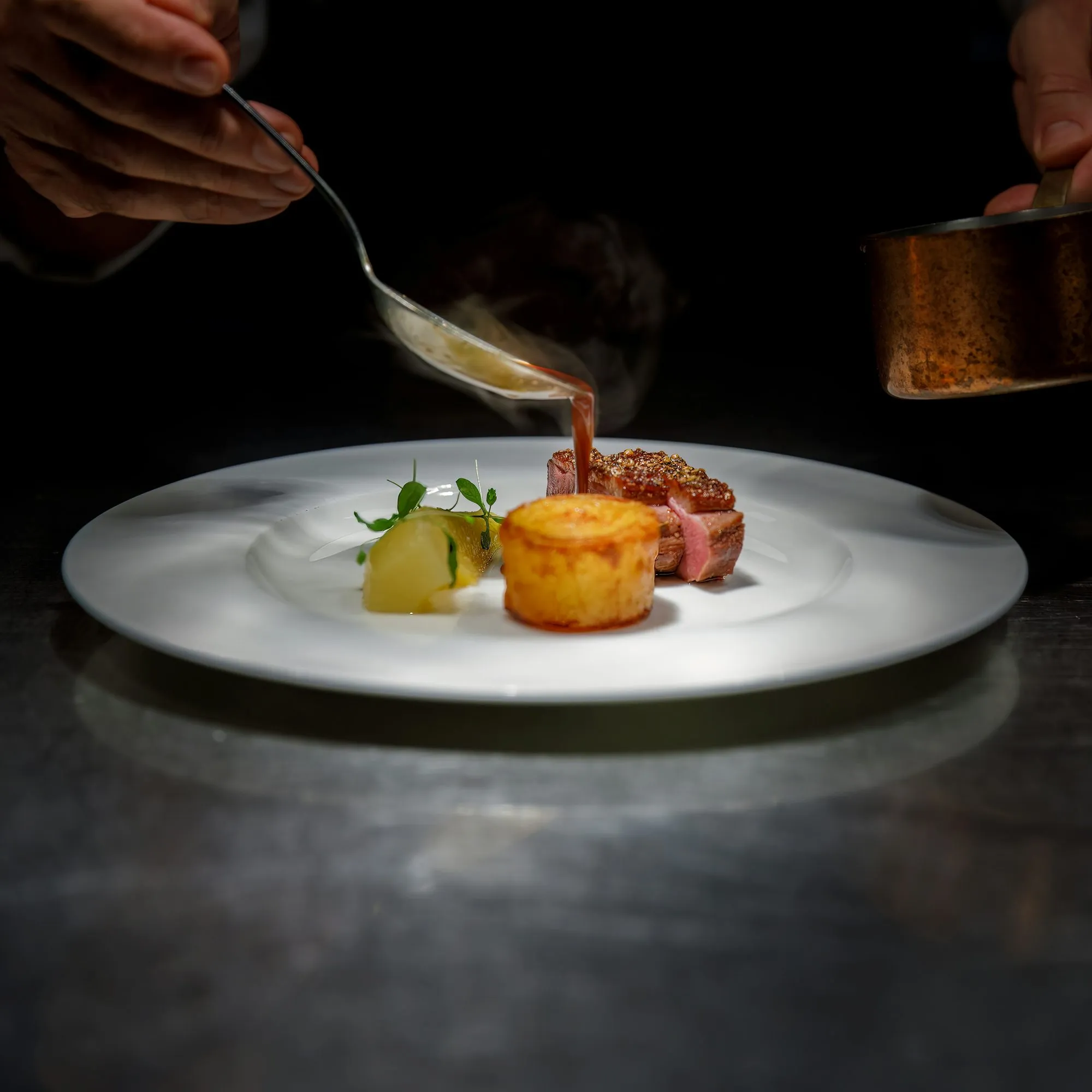 A chef spooning sauce over sliced duck breast with a potato cake on a white plate (stock photo)