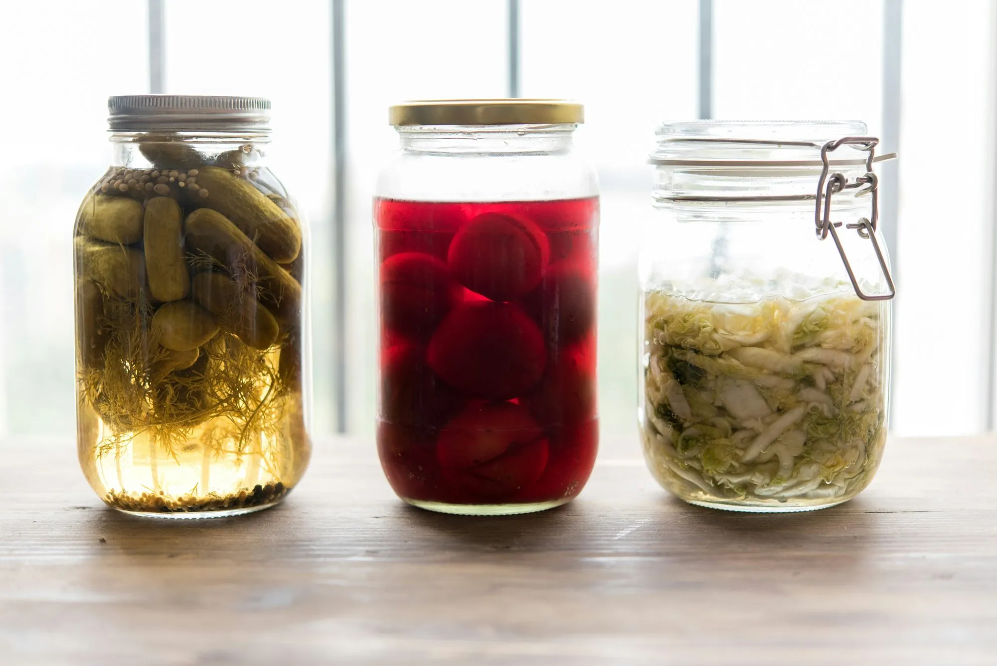 Three glass jars of pickled cucumbers, pickled beets and sauerkraut on a wooden table