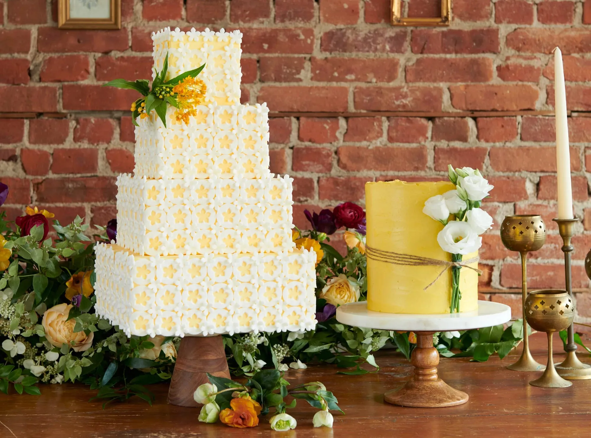 A four-tier square wedding cake covered in sugar flowers beside a yellow buttercream cake on a marble stand, against a brick wall