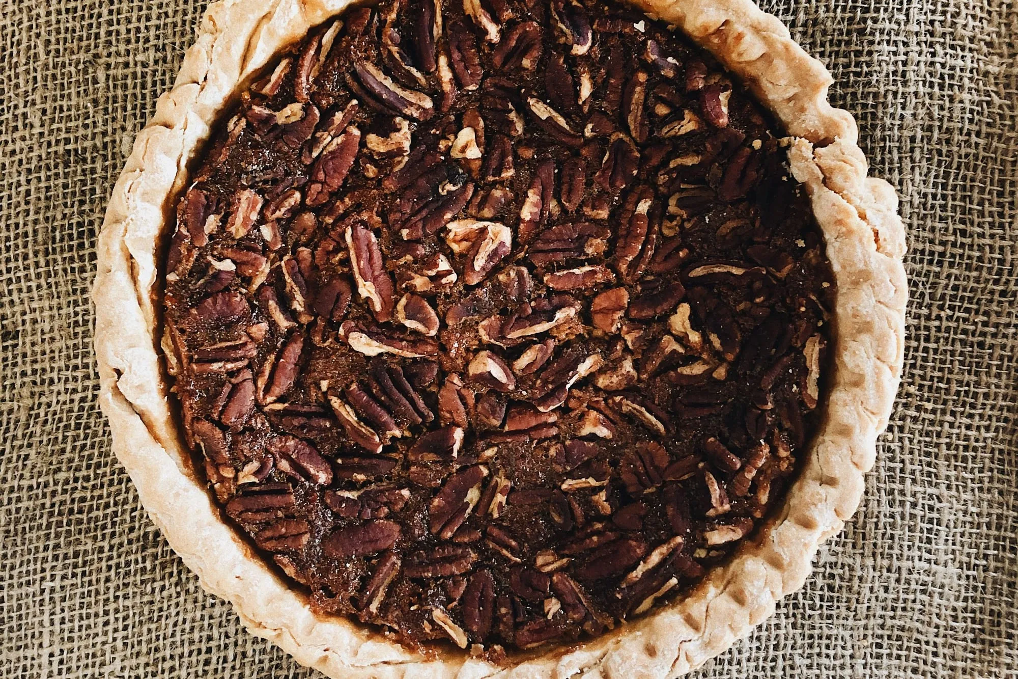 A whole pecan pie with a crimped crust on burlap, seen from above